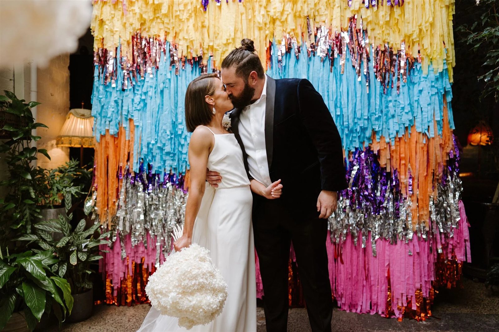 Newlyweds kissing in front of a colourful fringe backdrop